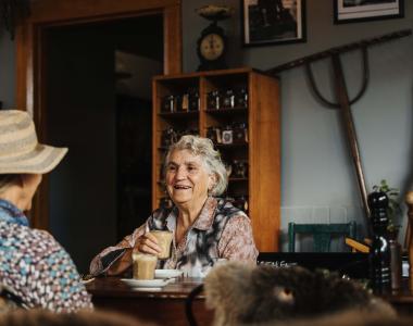 Lady with grey hair holding a coffee sits opposite a friend in a cafe with rustic decoration on walls